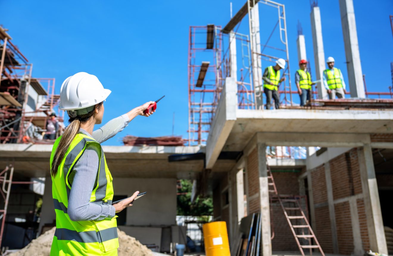 A woman in a hardhat and yellow vest points towards three construction workers in vests standing on a structure