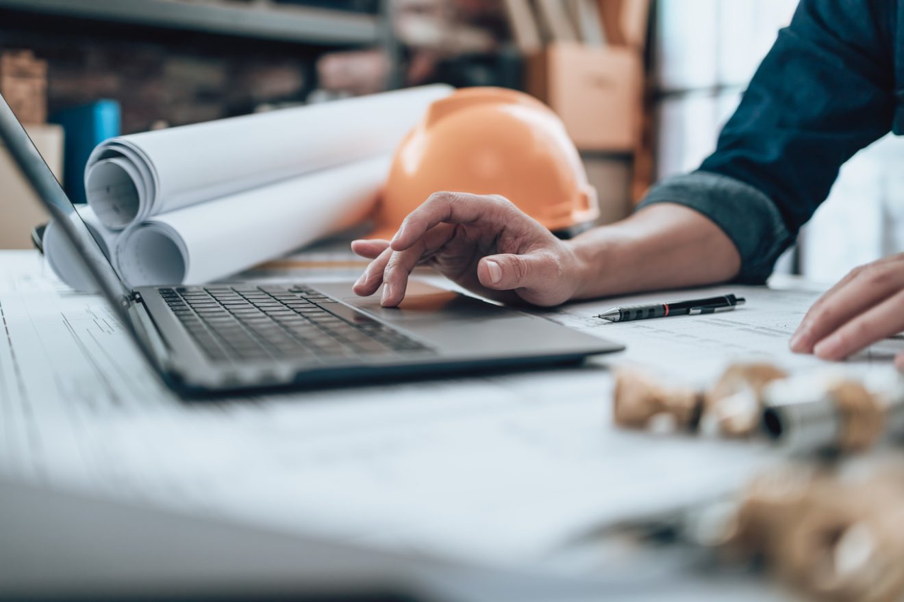 A person uses a laptop at a desk with rolled-up blueprints and a hardhat