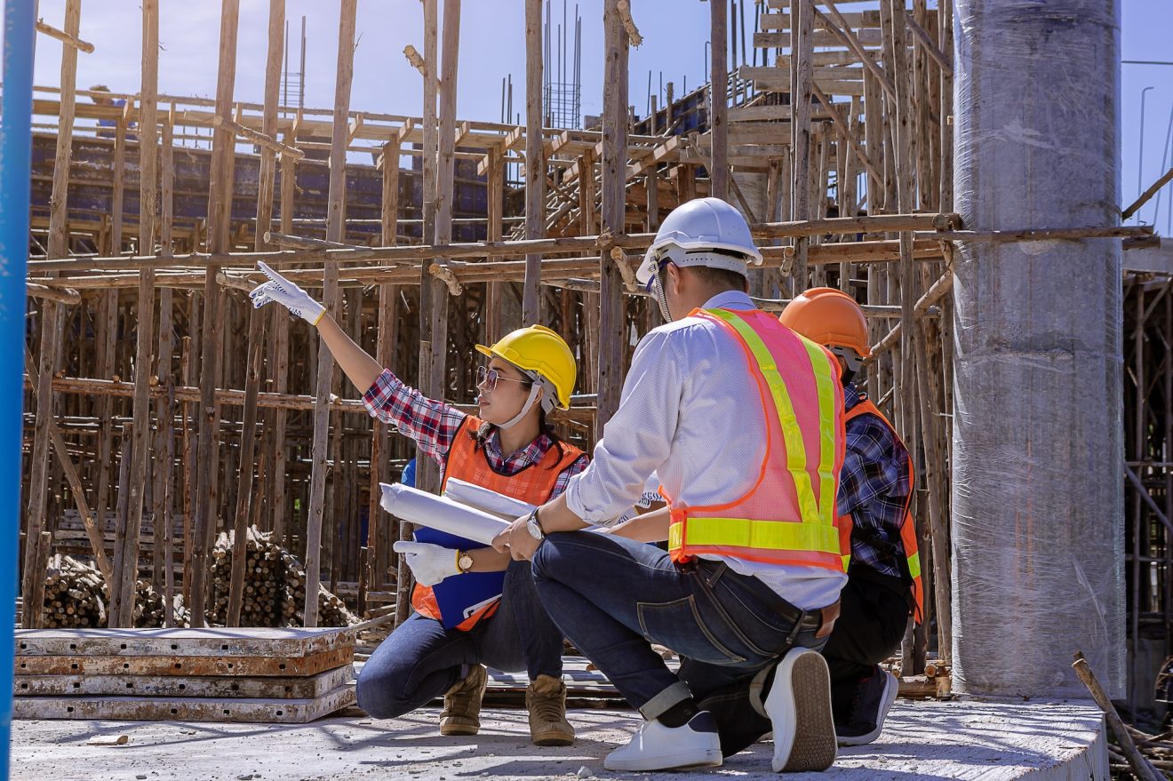 Three construction workers kneel at a jobsite and hold plans as one person points into the distance