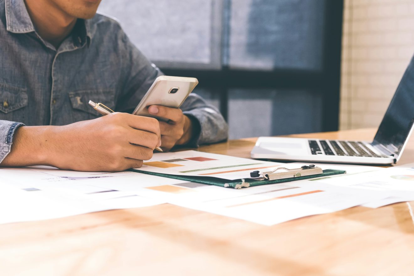 A person is sitting at their laptop, referencing their phone and physical notes in front of them