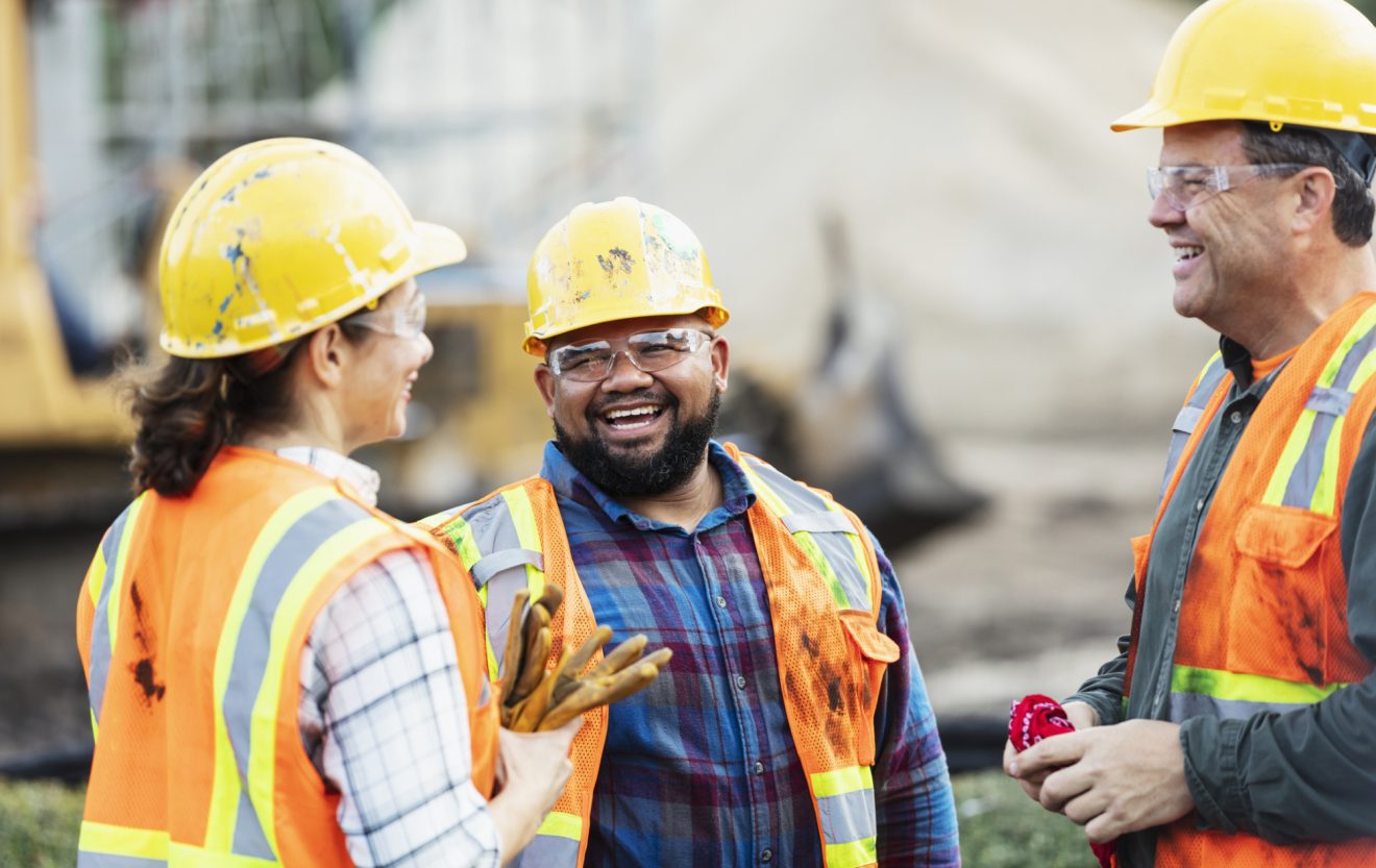 A group of three workers at a construction site wearing hard hats, safety glasses and reflective clothing, smiling and conversing