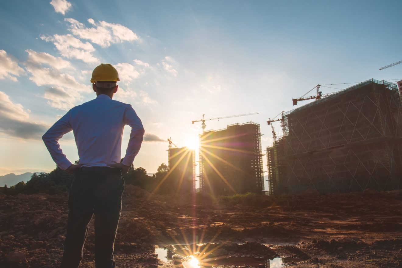 man in construction helmet looking out at silhouettes of building under construction in the distance