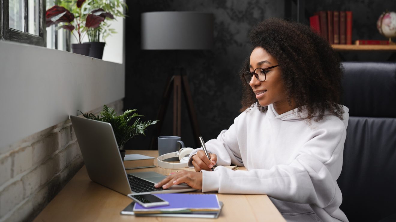 A woman takes notes in a notebook about something on her computer