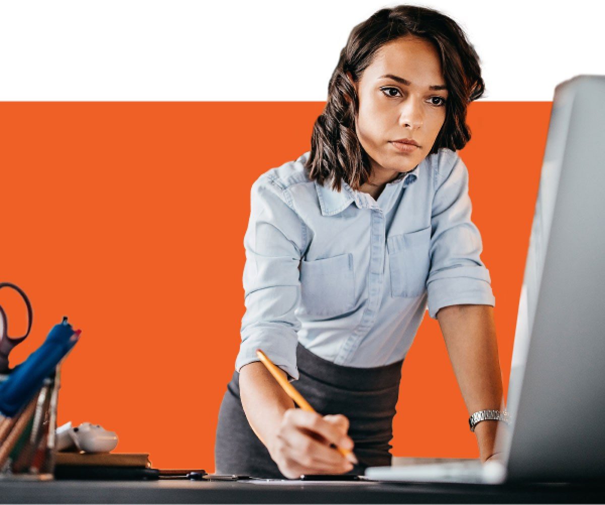 Woman writes at desk while looking at computer screen