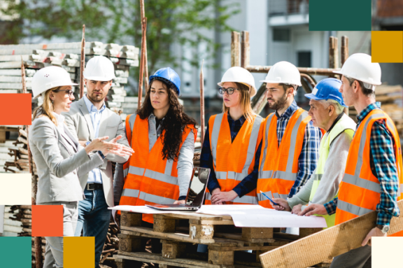 small group of men and women wearing hardhats and reflective vests listening to a woman in a gray suit and hardhat