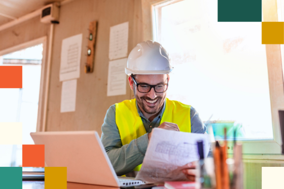 man in hardhat and reflective vest sitting at desk looking at laptop and planning papers
