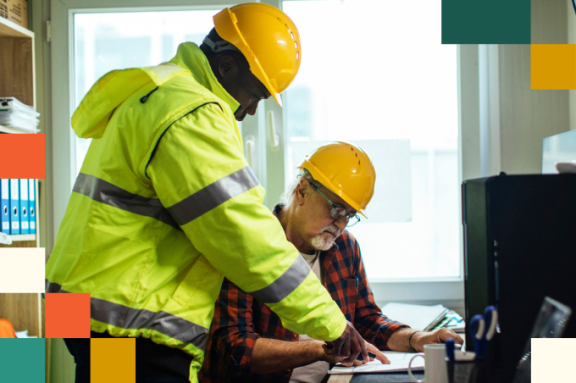 two men in hard hats reviewing a document in front of a desktop computer