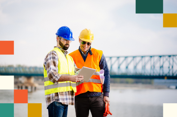 two men in hard hats and reflective vests look at tablet computer together