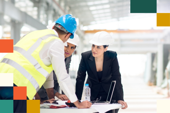 man in reflective vest and hard hat leaning over a desk speaking to man and woman in business attire and hard hats