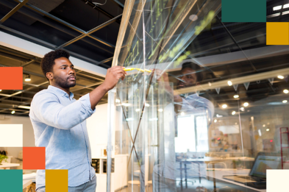 young man in blue dress shirt writes on transparent project board with a yellow marker
